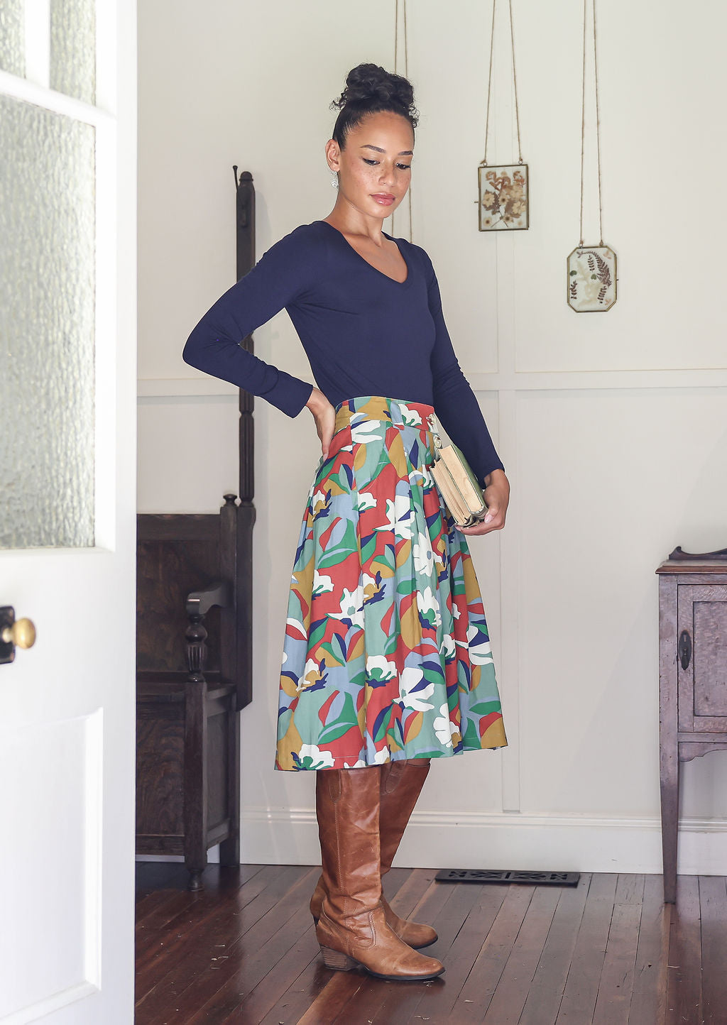Woman wearing a blue top and colorful skirt in a room with wooden floors and white walls.