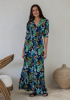 A woman modelling a navy blue rayon maxi dress with 3/4 sleeves, V-neckline, buttoned bodice and shirred waistband, in a colourful floral and peacock print