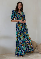 A woman modelling a navy blue rayon maxi dress with 3/4 sleeves, buttoned bodice and shirred waistband, in a colourful floral and peacock print