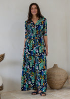 A woman modelling a 3/4 sleeved rayon maxi dress with a vibrant floral and peacock print in shades of navy blue, teal, green, and orange, standing in a minimalist room with a beige wall and woven decorative baskets.