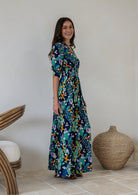A woman standing side on wearing a navy blue rayon maxi dress with 3/4 sleeves, buttoned bodice and a wide shirred waistband, in a colourful floral and peacock print