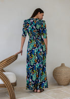 Back view of woman wearing a 3/4 sleeved rayon maxi dress with a vibrant floral and peacock print in shades of teal, green, blue, orange, and purple on a dark navy base.