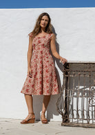 Model poses in a knee length 100% cotton dress in cream based floral print with scoop neck, pockets and cap sleeves