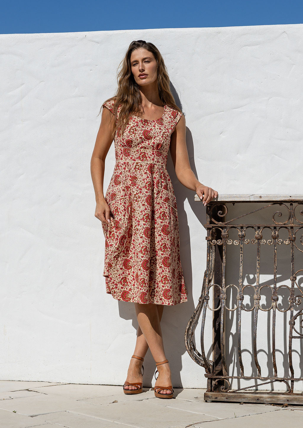 Woman in a floral dress standing against a white wall with a metal gate.