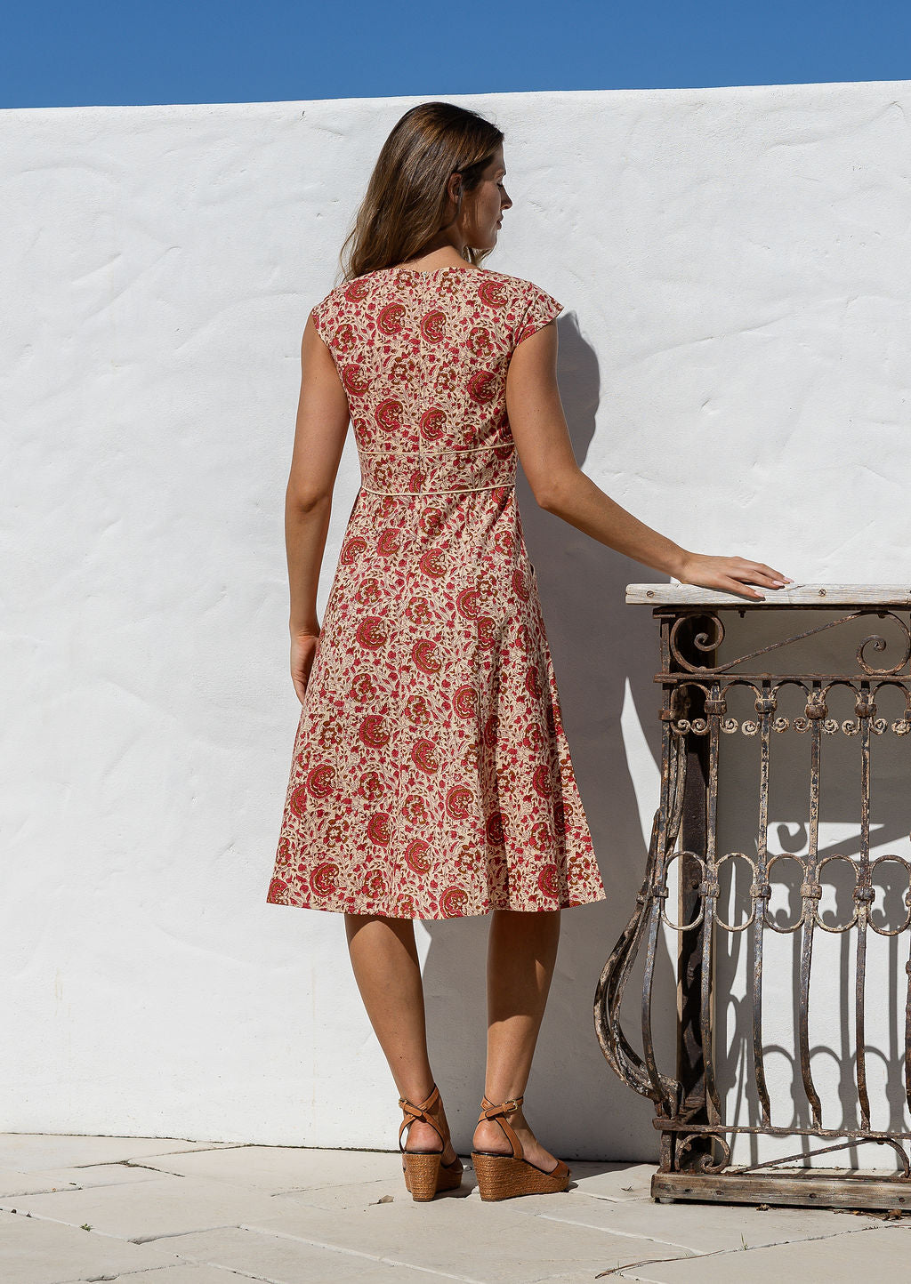 Woman standing backwards wearing a cream based floral print dress with scoop neck, cap sleeves and piped detailing at the waist and pockets.