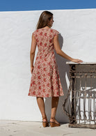 Woman standing backwards wearing a cream based floral print dress with scoop neck, cap sleeves and piped detailing at the waist and pockets.