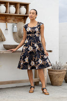 Woman wearing a floral cotton dress with cap sleeves, piped details standing in a rustic kitchen setting.