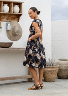 Model standing backwards of white wall wearing a retro style 100 percent cotton dress black based floral print. 