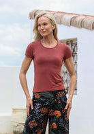Woman wearing a terracotta fitted t-shirt and floral pants standing against a white wall