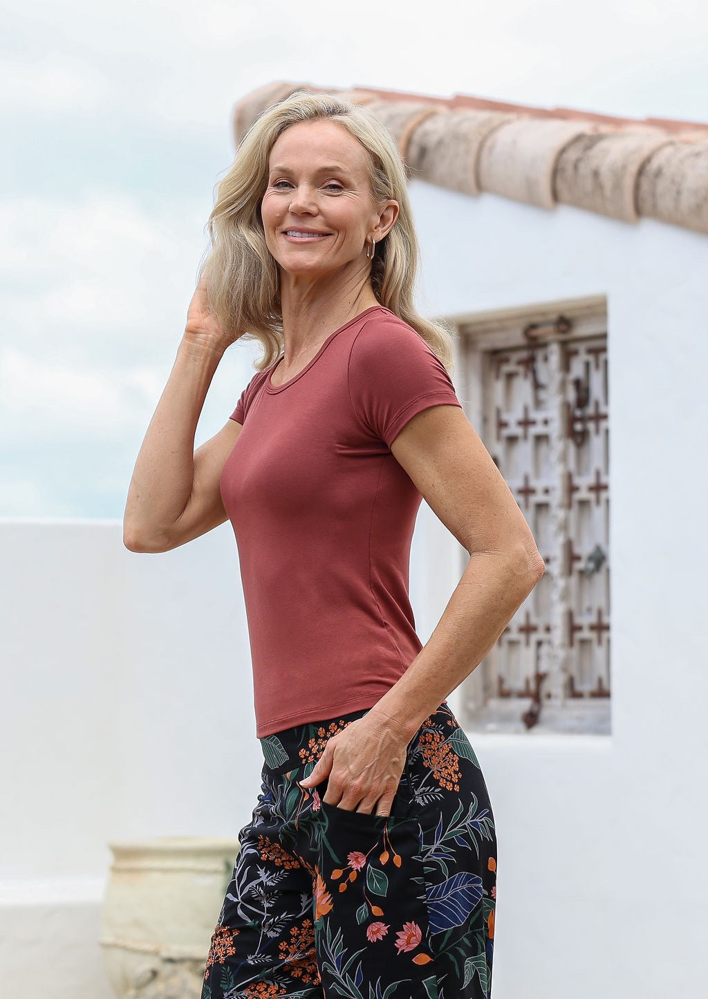 Woman wearing a terracotta shirt and floral pants standing outdoors with a house in the background