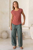 Woman wearing a 100% rayon terracotta shell t-shirt and green pants standing in a room with white walls and decorative vases.