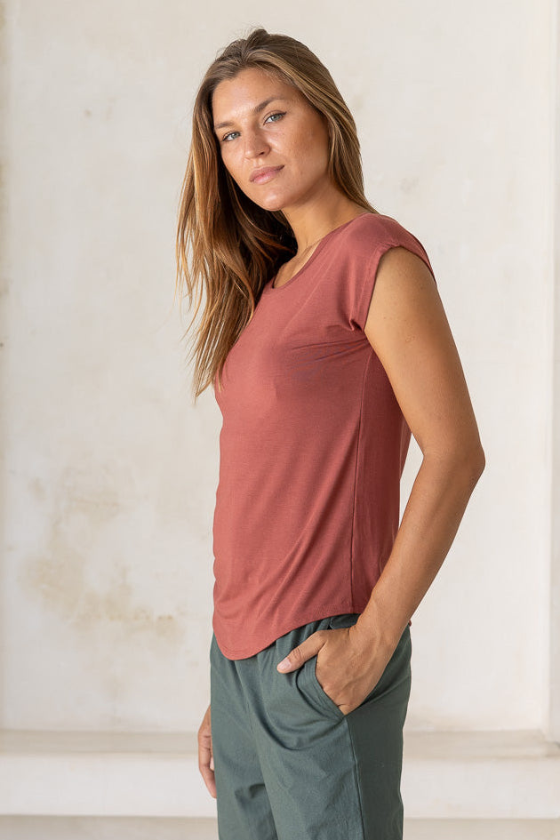 Woman stands sideways wearing a 100% rayon terracotta shell t-shirt and green pants standing in a room with white walls and light-colored floor.