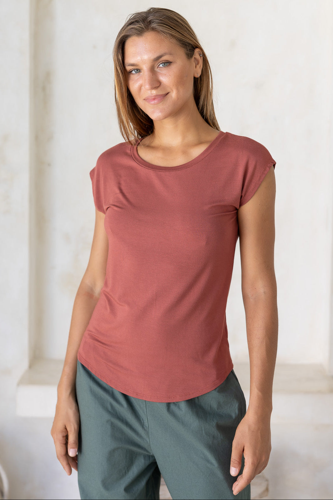 Woman wearing a 100% rayon terracotta shell t-shirt and green cotton pants against a white wall.