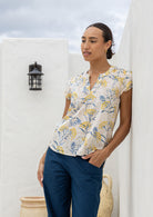 Woman wearing a neutral based floral  blouse and blue pants standing against a white wall.