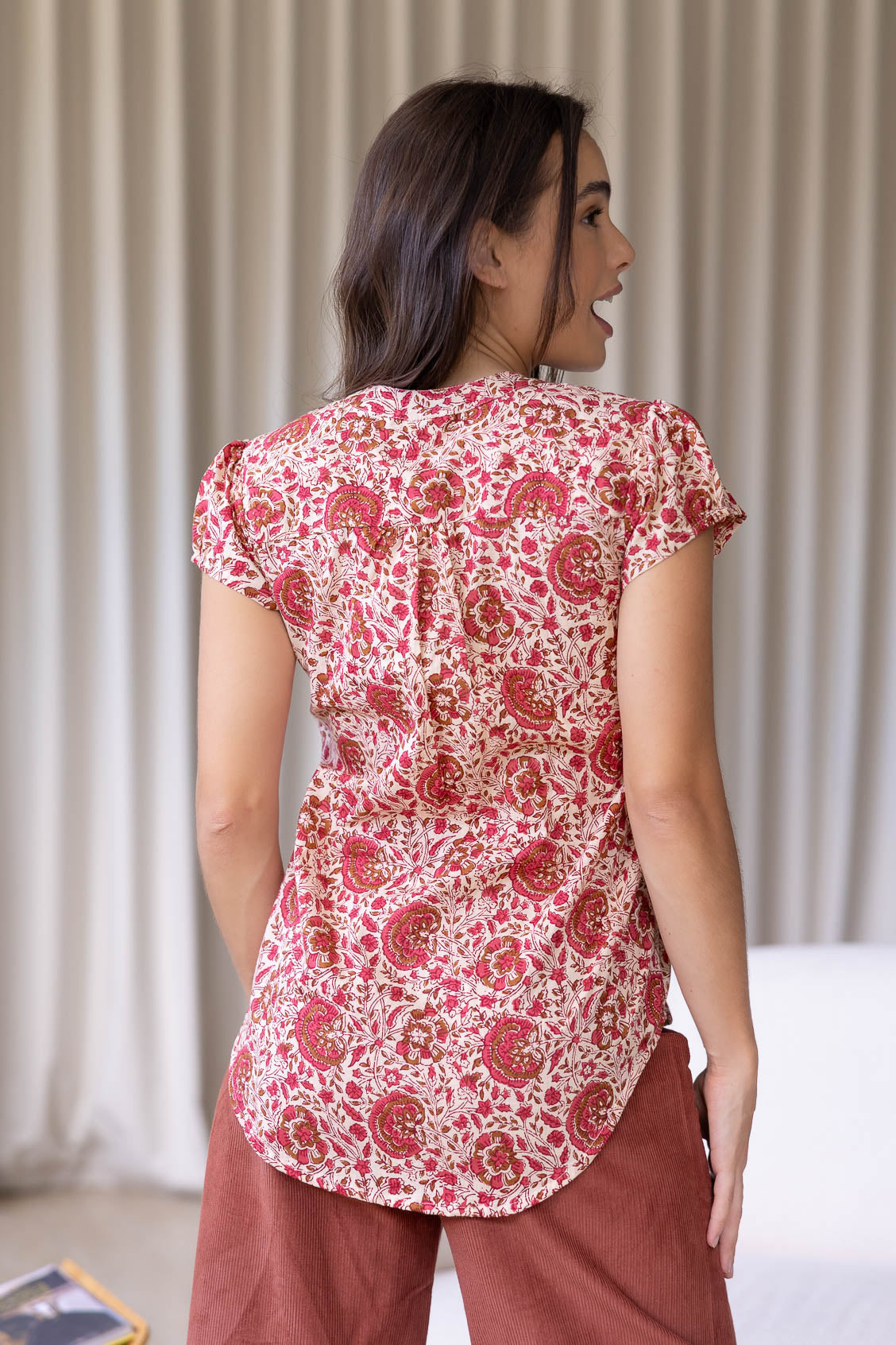 Woman stands backwards wearing a 100% cotton warm gentle cream based floral blouse and plain pants.