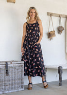Woman wearing a 100% cotton button bodice sleeveless dress in black based floral print with one hand in pocket