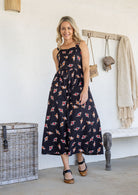 Woman wearing a 100% cotton dress in black based floral print paired with tan sandals.
