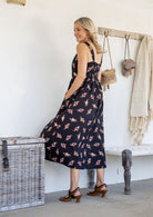 Woman wearing a 100% cotton button bodice sleeveless dress in white based floral print with both hand in pockets