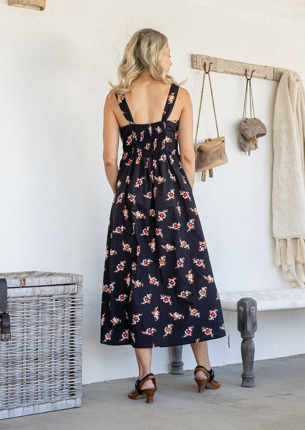 Woman standing backwards wearing a 100% cotton button bodice sleeveless dress in black based floral print with elasticated back and front pockets