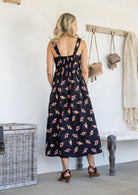Woman standing backwards wearing a 100% cotton button bodice sleeveless dress in black based floral print with elasticated back and front pockets