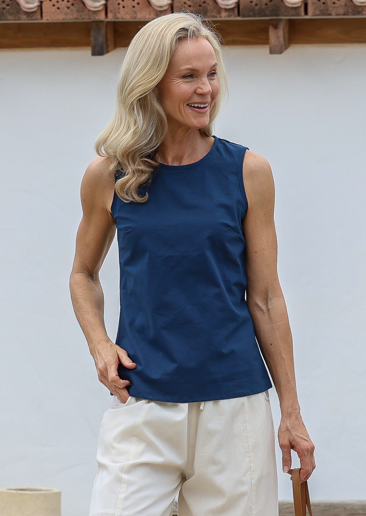 Woman wearing a blue sleeveless, relaxed fit cotton top with round neckline and piped detailing, paired with white cotton pants 