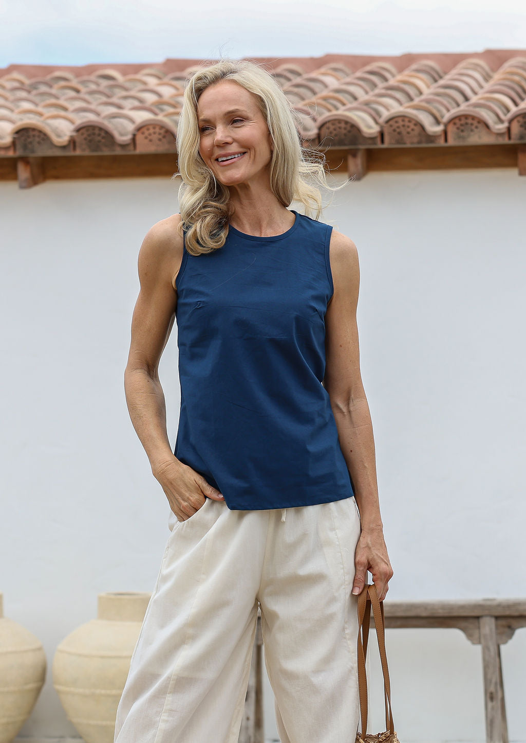 Woman wearing a blue sleeveless cotton top with side bust darts and off white pants standing against a white wall with a terracotta roof.