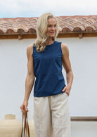 Woman stands with one hand in pants pocket wearing a 100% cotton relaxed fit, sleeveless sea blue top with piped detailing at the neckline and armholes 