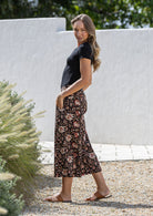 Woman stands sideways both hand in pockets wearing a black based floral print wide leg, 3/4 length cotton pants with side zipper 