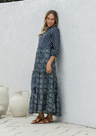 Woman wearing a 100% cotton tiered maxi dress in navy blue based floral print against a white wall with white vases in the foreground.