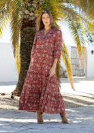 Woman poses in red based floral cotton maxi dress with buttoned bodice, 3/4 sleeves and flowy three tiered maxi skirt