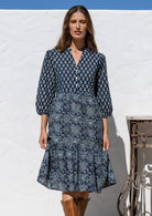 Woman wearing a blue patterned dress and brown boots standing against a white wall.