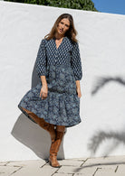 Woman wearing a blue floral dress with brown boots against a white wall.