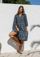 Woman in a floral dress and brown boots standing against a white wall.