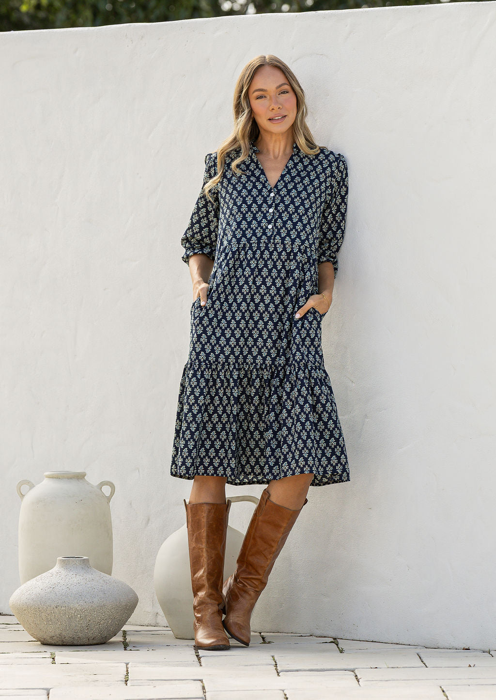 Woman standing in cotton tiered over the knee dress with 3/4 sleeves, buttoned bodice, V-neckline and hands in pockets