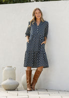 Woman standing in cotton tiered over the knee dress with 3/4 sleeves, buttoned bodice, V-neckline and hands in pockets