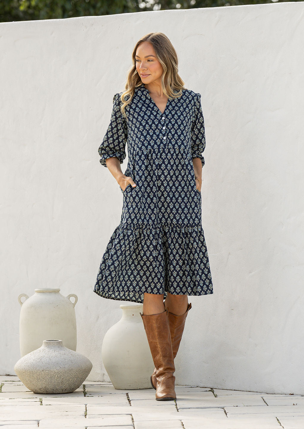 Woman wearing a navy blue based floral dress and brown boots standing in front of a white wall.