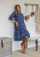 Woman wearing a blue patterned dress and brown boots standing against a white wall.