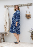 Woman stands sideways in cotton tiered over the knee dress with 3/4 sleeves, buttoned bodice and V-neckline