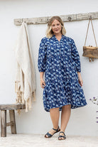 Woman wearing a blue patterned dress and brown boots standing against a white wall.
