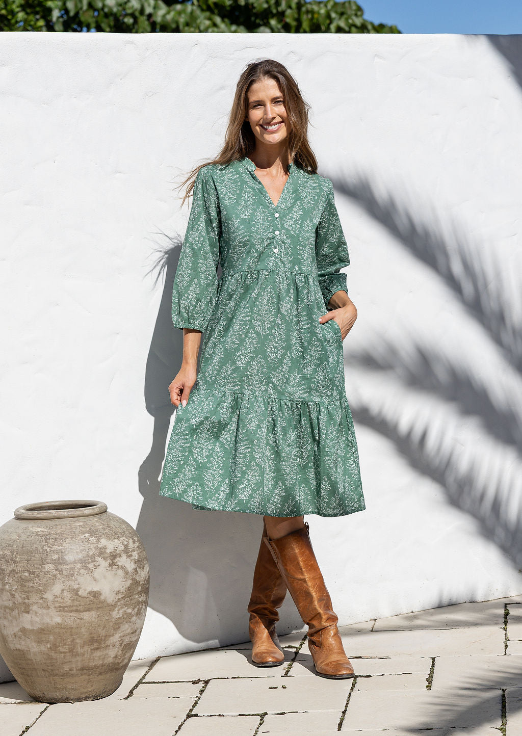 Woman wearing a sage green based floral dress and brown boots standing in front of a white wall.