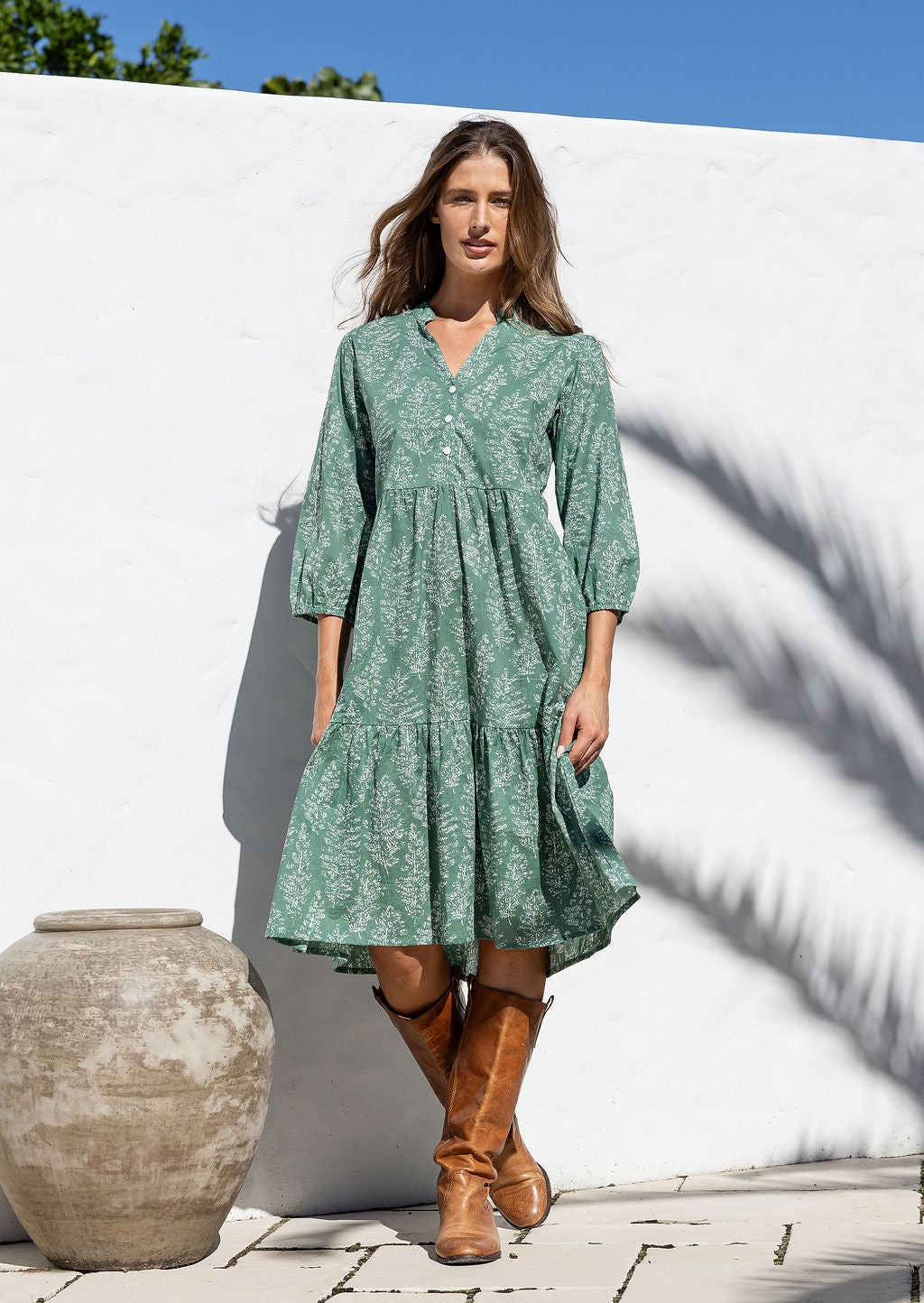 Woman in a floral dress with pockets and brown boots standing in front of a white wall.