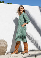 Model stands side on in cotton over the knee length relaxed fit dress with 3/4 sleeves, paired with brown boots. 
