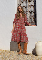 Woman wearing a red based floral dress and brown boots standing in front of a decorative wooden door.