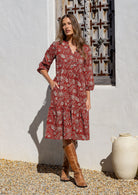 Woman in a floral dress and brown boots standing in front of a rustic wooden door with one hand in pocket. 