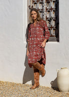 Woman in a red floral dress and brown boots standing against a white wall with a decorative metal gate.