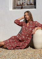 Woman in a red floral dress sitting against a white wall with a window.