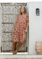 Woman in a floral dress and brown boots standing in front of a rustic wooden door.
