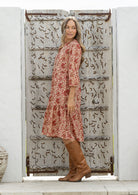 Woman in a red floral dress and brown boots standing in front of a decorative wooden door.