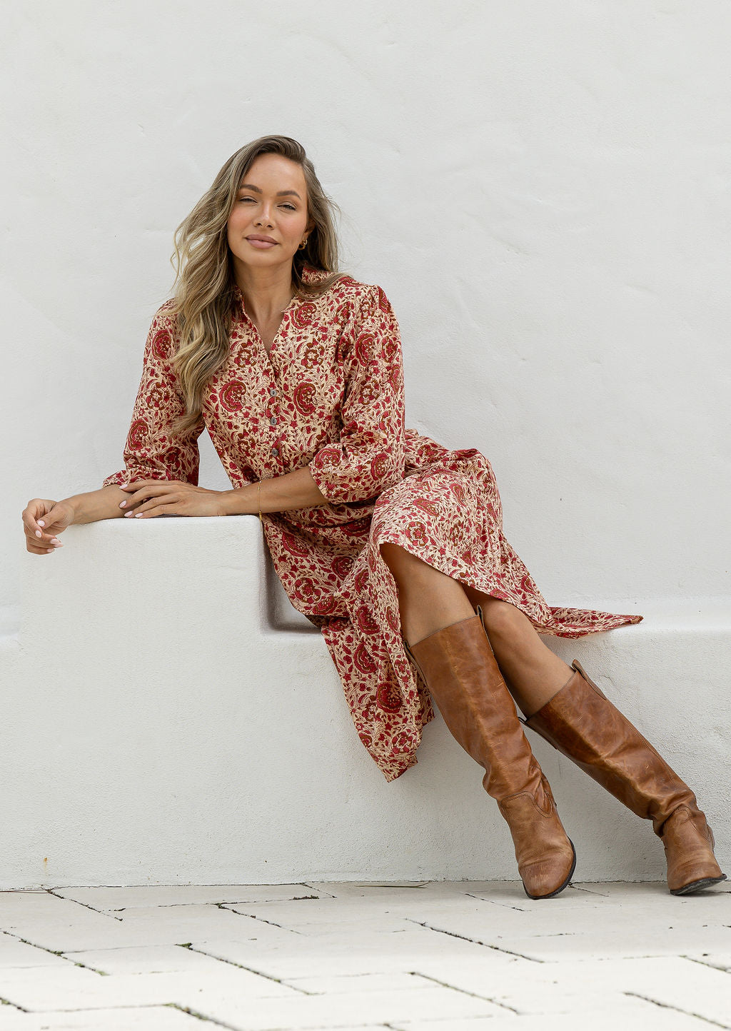 Woman in a floral dress and brown knee-high boots sitting on a white ledge.