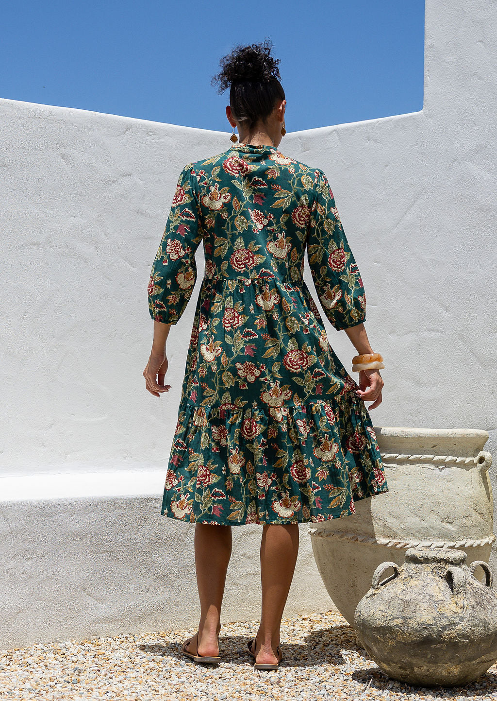 Model stands backwards in cotton tiered over the knee floral print dress, holding the skirt out, paired with tan sandals